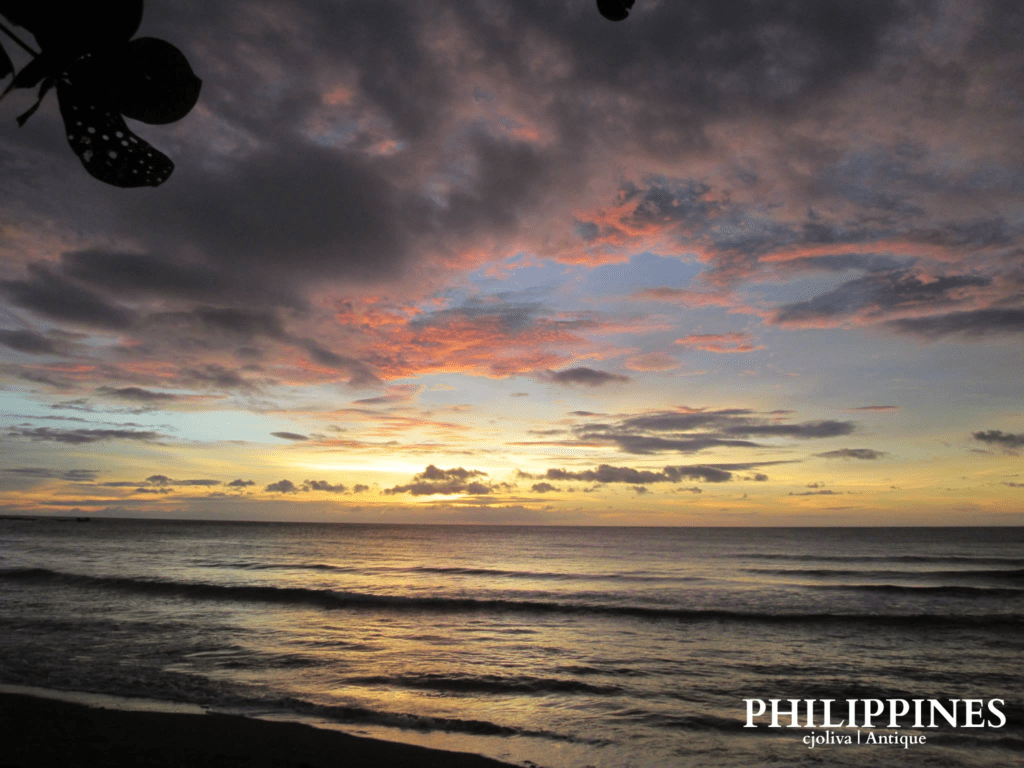 Scenic sunset over the ocean in Antique, Philippines, with waves rolling toward shore, dramatic clouds illuminated in orange, pink, and purple hues, and silhouetted leaves framing the view.