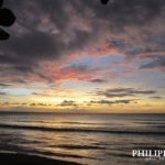 Scenic sunset over the ocean in Antique, Philippines, with waves rolling toward shore, dramatic clouds illuminated in orange, pink, and purple hues, and silhouetted leaves framing the view.