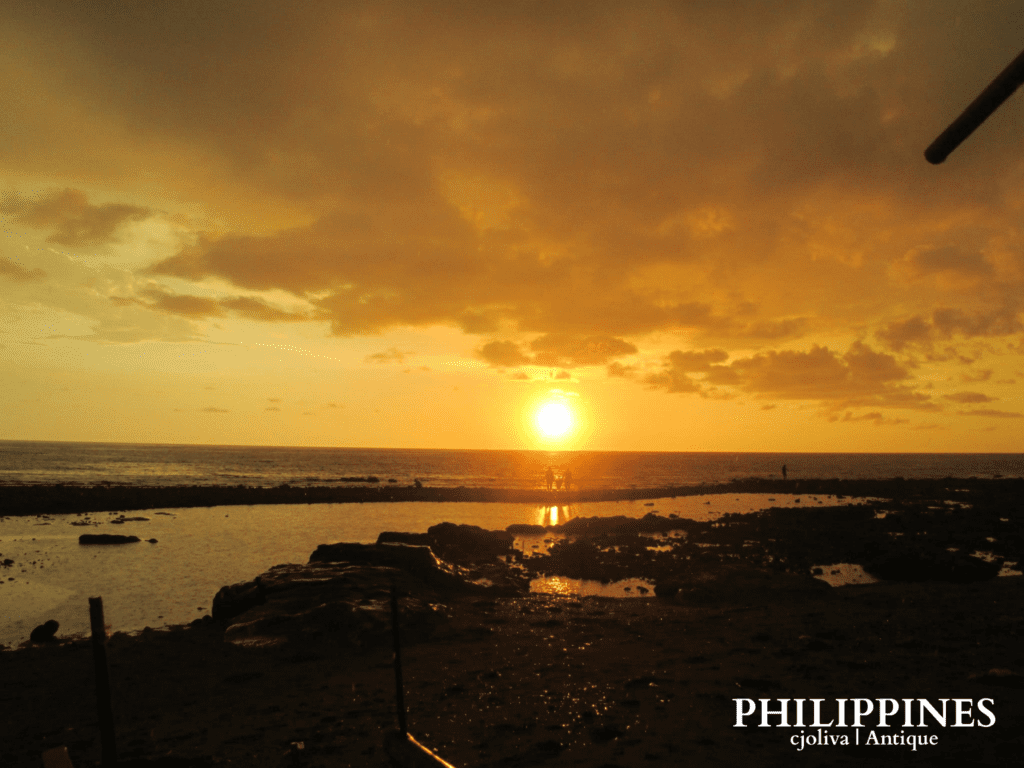 Scenic sunset over rocky shoreline in Antique, Philippines, with golden sky, ocean reflections, silhouetted figures, and dramatic clouds, taken in 2018