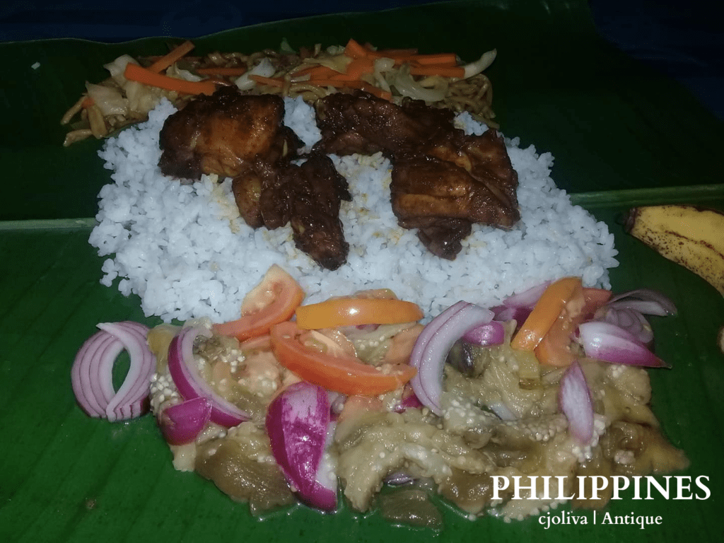 A traditional Filipino meal served on a banana leaf in Antique, Philippines, with rice, adobo-style meat, sautéed vegetables, ensaladang talong, and a banana, taken in 2018