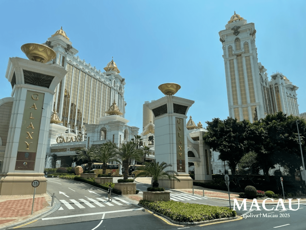A grand entrance to the Galaxy Macau resort, featuring towering white and gold skyscrapers with cupolas, ornate pillars, and lush palm trees under a bright blue sky.
