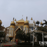 A view of the Arabian-themed palace at Leofoo Village Theme Park in Taiwan, featuring golden domes, intricate Middle Eastern architectural patterns, palm trees, and an elevated track ride against a cloudy sky.