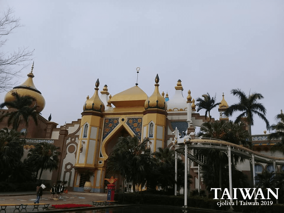 A view of the Arabian-themed palace at Leofoo Village Theme Park in Taiwan, featuring golden domes, intricate Middle Eastern architectural patterns, palm trees, and an elevated track ride against a cloudy sky.