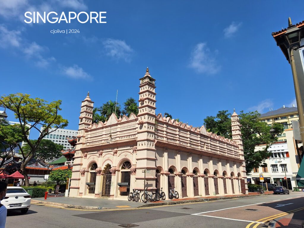 The Nagore Dargah mosque in Singapore has Indo-Islamic architecture, a pink and cream façade, ornate towers, and bicycles parked in front under a clear blue sky.
