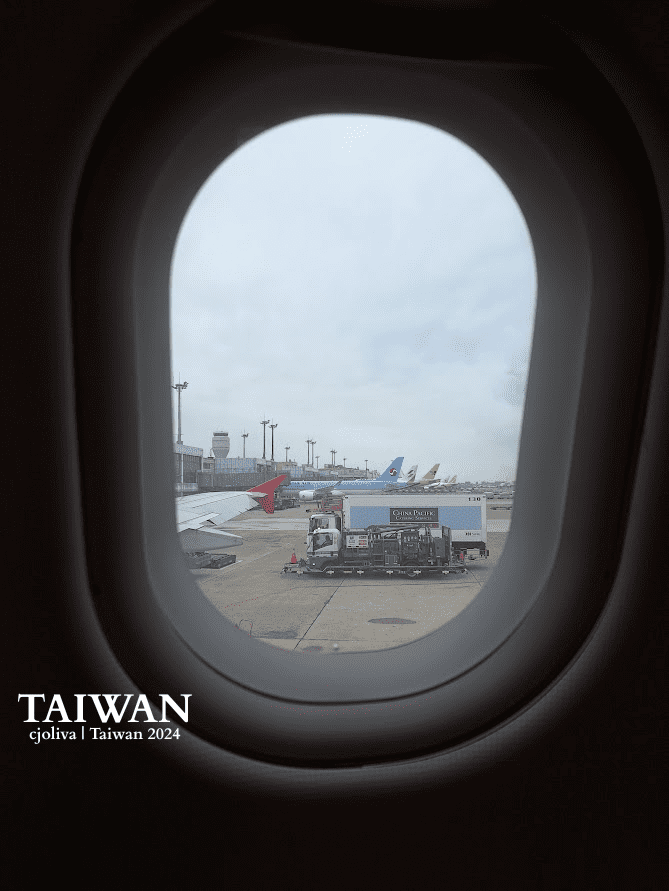 View from an airplane window of an airport tarmac with aircraft tails from multiple airlines, a catering truck, and an overcast sky.