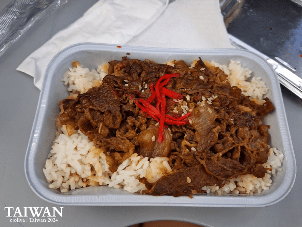 Taiwanese beef rice bowl with marinated beef slices, onions, sesame seeds, red pepper garnish, and white rice in a rectangular container.