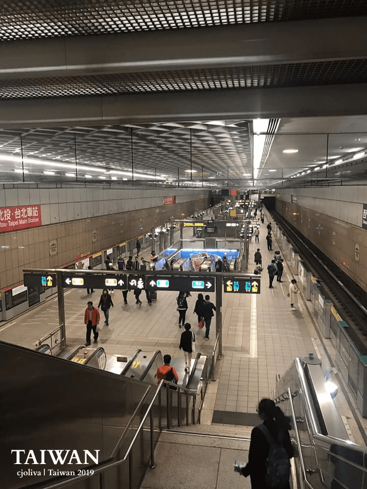 Underground view of Taipei Main Station in Taiwan with tiled walls, grid‑patterned ceiling, directional signage for exits and transfers, escalators, and people walking through the well‑lit platform area.