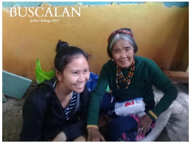 An elderly indigenous woman wearing traditional beads and headwear is sitting beside a smiling young woman against a yellow wall background in Buscalan.