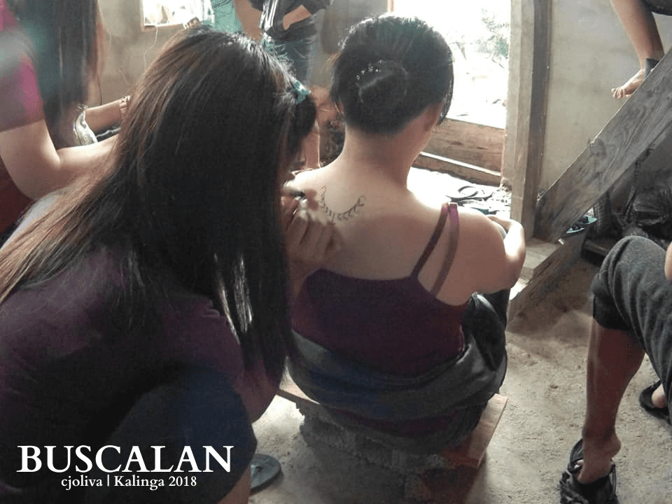 A woman receiving a traditional hand-tapped tattoo on her upper back in Buscalan, Kalinga, with another person working carefully on the tattoo design.
