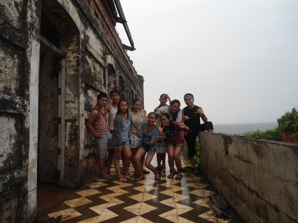 A group of nine friends poses and smiles on the checkered tile balcony of the weathered, historic Capones Lighthouse in Anawangin, Zambales. The brick walls show aging and character, with the sea visible in the distance under an overcast sky.