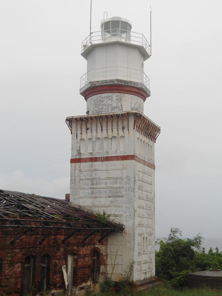 Historic Capones Lighthouse with weathered white tower, red bands, and attached brick ruins surrounded by greenery