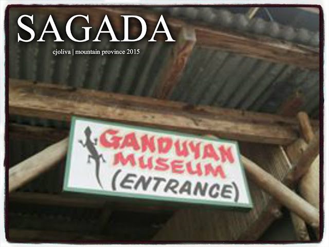 Wooden signboard for Ganduyan Museum entrance in Sagada, Mountain Province, mounted on a wooden frame with a corrugated roof and lizard illustration.