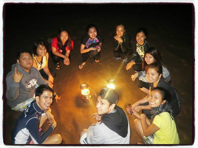 A group of friends sitting in a circle on the ground at night, illuminated by two lanterns in the center, smiling and enjoying each other's company.