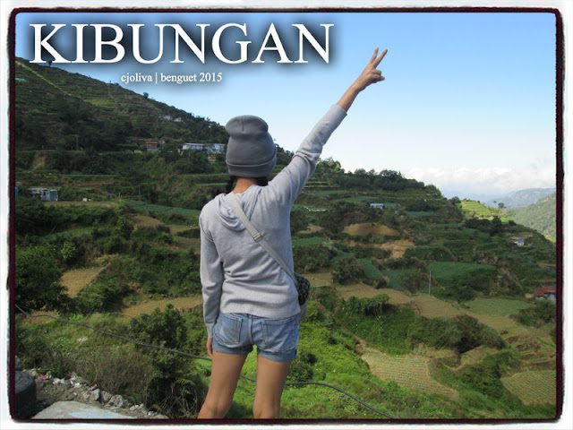 Person raising a peace sign on a hillside overlooking lush green terraces, scattered houses, and mountain ranges in Kibungan, Benguet.