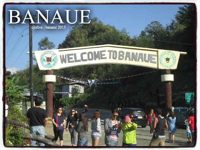 Tourists gathered under the "Welcome to Banaue" archway sign at the entrance to Banaue, surrounded by greenery and vehicles in the background.