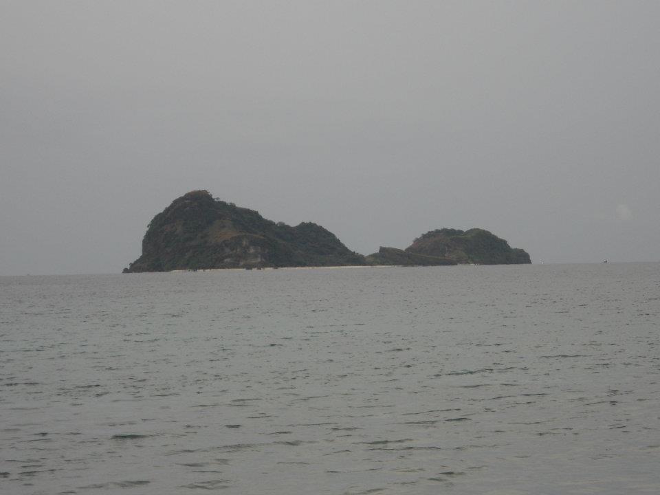 Small rugged island with dense vegetation and hilly terrain surrounded by calm ocean waters under an overcast sky