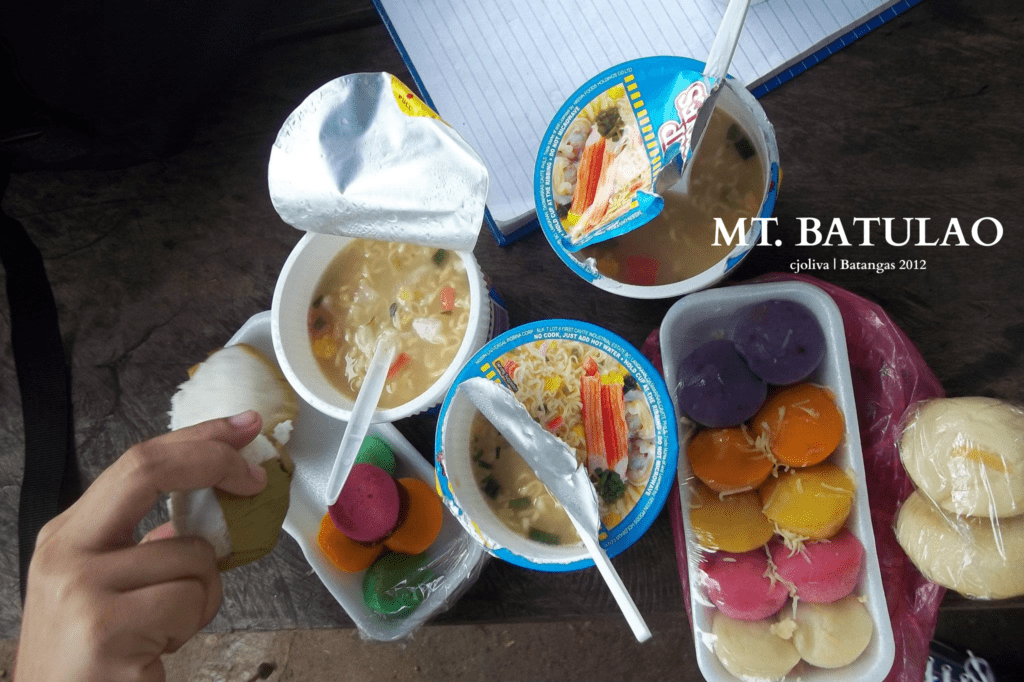 Instant noodles cups and colorful Filipino kakanin snacks on a table, with a hand holding a rice cake, during Mt. Batulao hike
