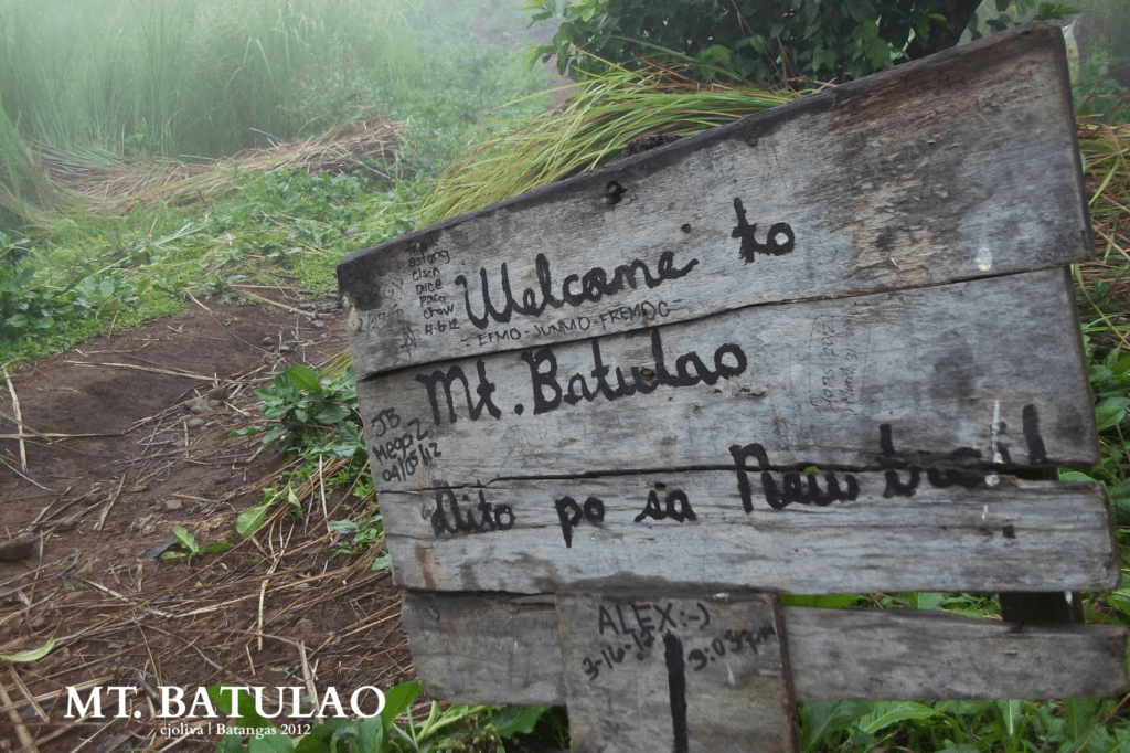 A weathered wooden welcome sign for Mt. Batulao New Trail in Nasugbu, Batangas, surrounded by green foliage and mountain mist.