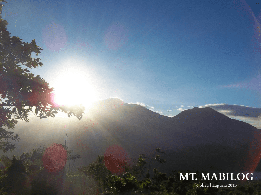 A bright sunrise casting rays over Mt. Mabilog with silhouetted mountain peaks and trees in the foreground, Laguna, 2013.