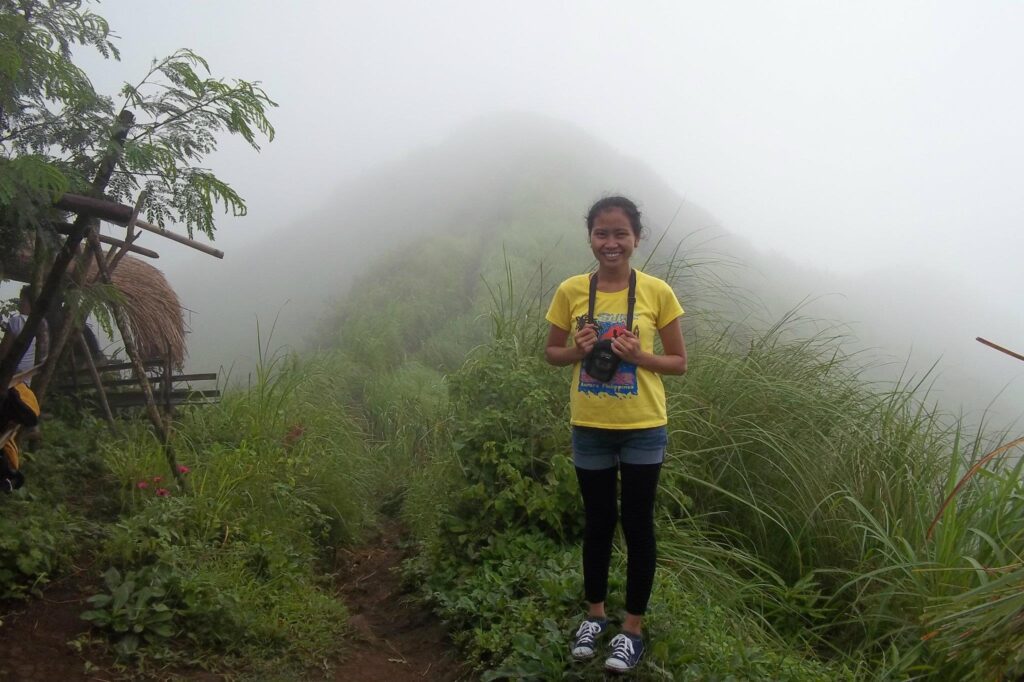 Woman in yellow shirt and black leggings standing on a foggy mountain trail holding a camera, surrounded by green plants and mist