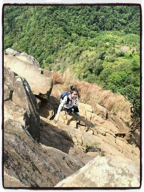 A female hiker carefully climbing the steep, rocky side of the Pico de Loro monolith with a lush forest background.