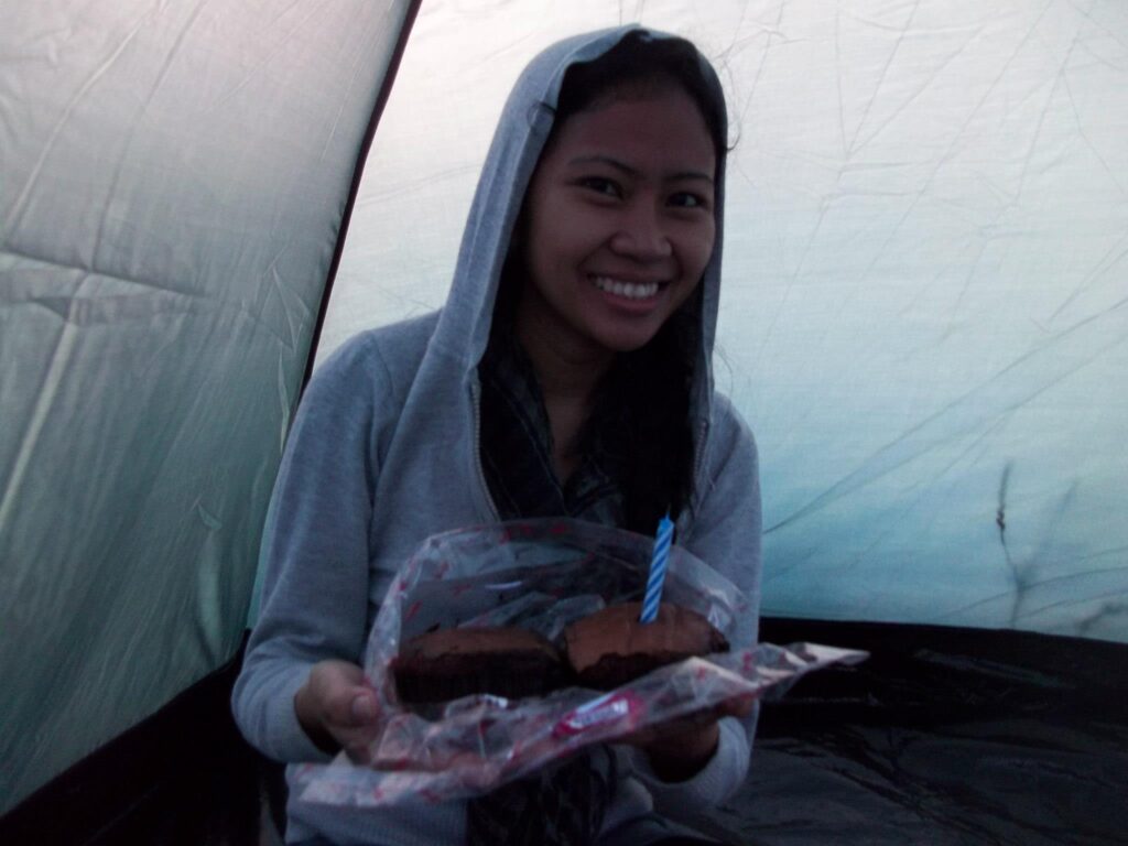 A smiling female hiker in a gray hoodie is sitting inside a tent, holding a chocolate cupcake with a single blue candle during a birthday climb.