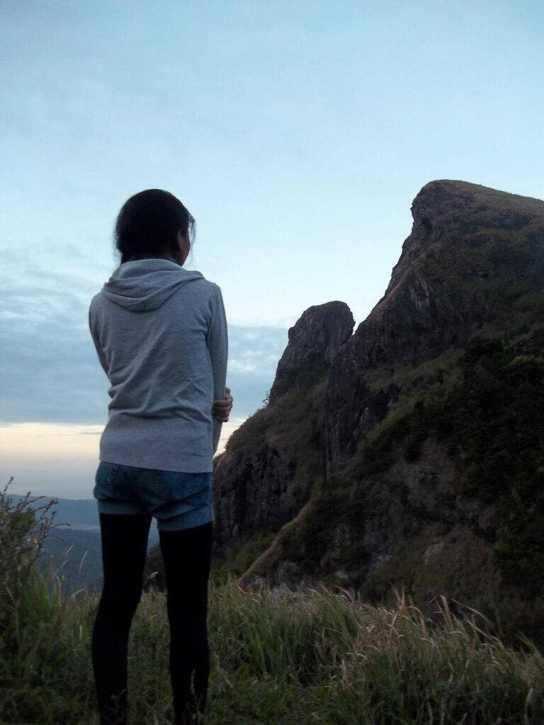 A female hiker from behind, wearing a gray hoodie, looking out over a view of the Pico de Loro monolith and mountains from the campsite at dawn.