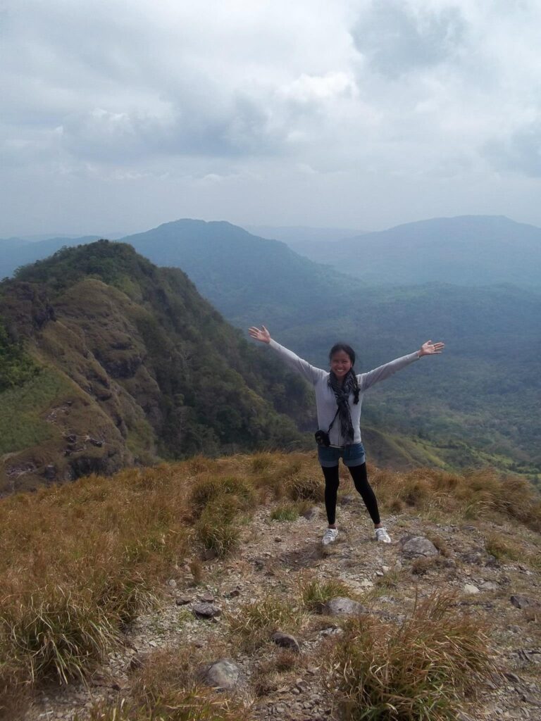 A smiling female hiker in a gray hoodie with arms wide open, celebrating her birthday solo climb on the grassy summit of Pico de Loro with a background of rolling green mountains.