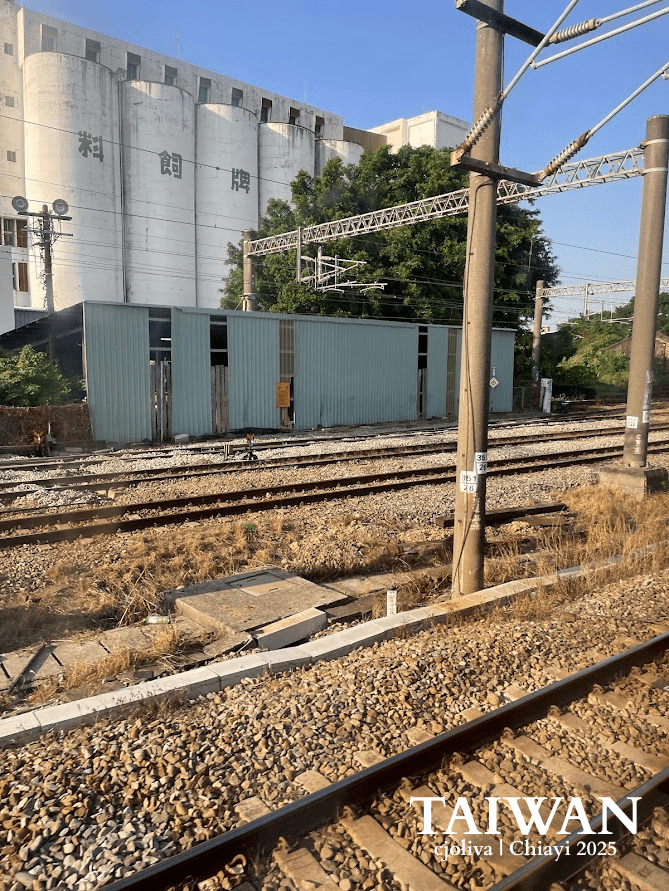 View from a moving train window of large industrial grain silos and feed mills next to the railway tracks near Chiayi, Taiwan, under a bright sky.