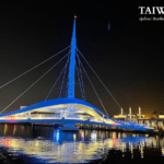 Night view of the Great Harbor Bridge (Dagang Bridge) in Kaohsiung, Taiwan. The white cable-stayed bridge is illuminated in blue, reflecting on the water near Pier-2.