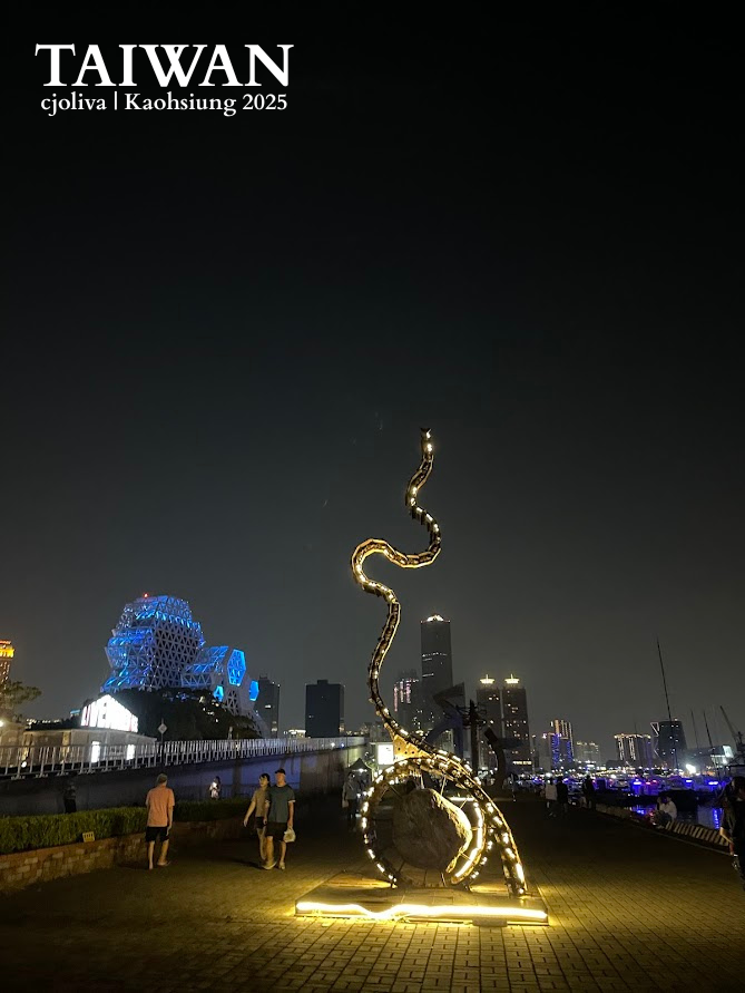 A winding illuminated outdoor sculpture at Kaohsiung Harbor at night, with the blue Kaohsiung Music Center and city skyline in the background.