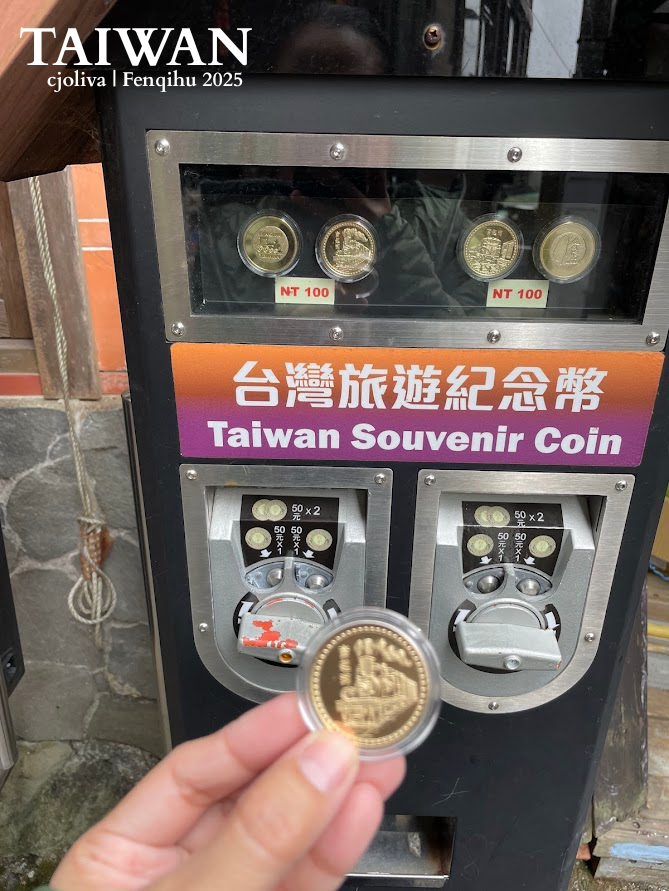 A hand holding a gold-colored souvenir coin in a plastic case in front of a "Taiwan Souvenir Coin" vending machine in Fenqihu, featuring train-themed designs.