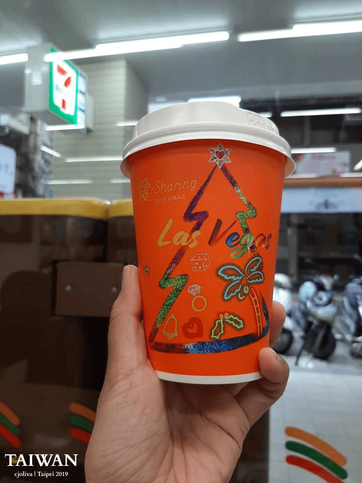 A close-up of a hand holding a bright orange 2019 Christmas edition coffee cup from 7-Eleven Taiwan. The cup features a "Las Vegas" themed Christmas tree design with palm trees and neon-style icons, set against a blurred convenience store background.