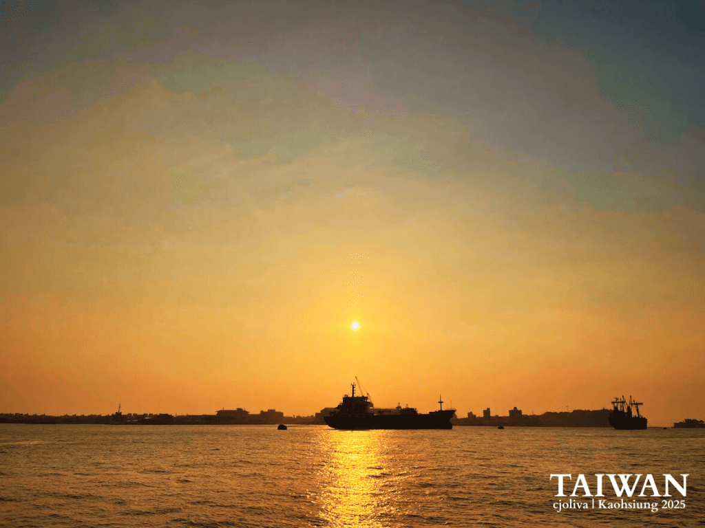 A wide-angle shot of a cargo ship silhouetted against a vibrant orange and yellow sunset in the Kaohsiung harbor, with the city skyline visible on the horizon.