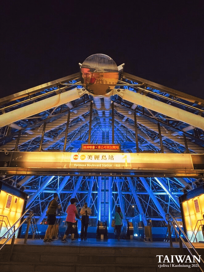 The striking glass and steel pyramid entrance of Formosa Boulevard Station in Kaohsiung at night, illuminated with glowing blue and yellow lights as people enter the station.