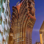 A low-angle nighttime shot of the Grand Lisboa hotel in Macau. The skyscraper's unique lotus-shaped architecture is illuminated in bright gold lights against a deep blue dusk sky, framed by older apartment buildings in the foreground.