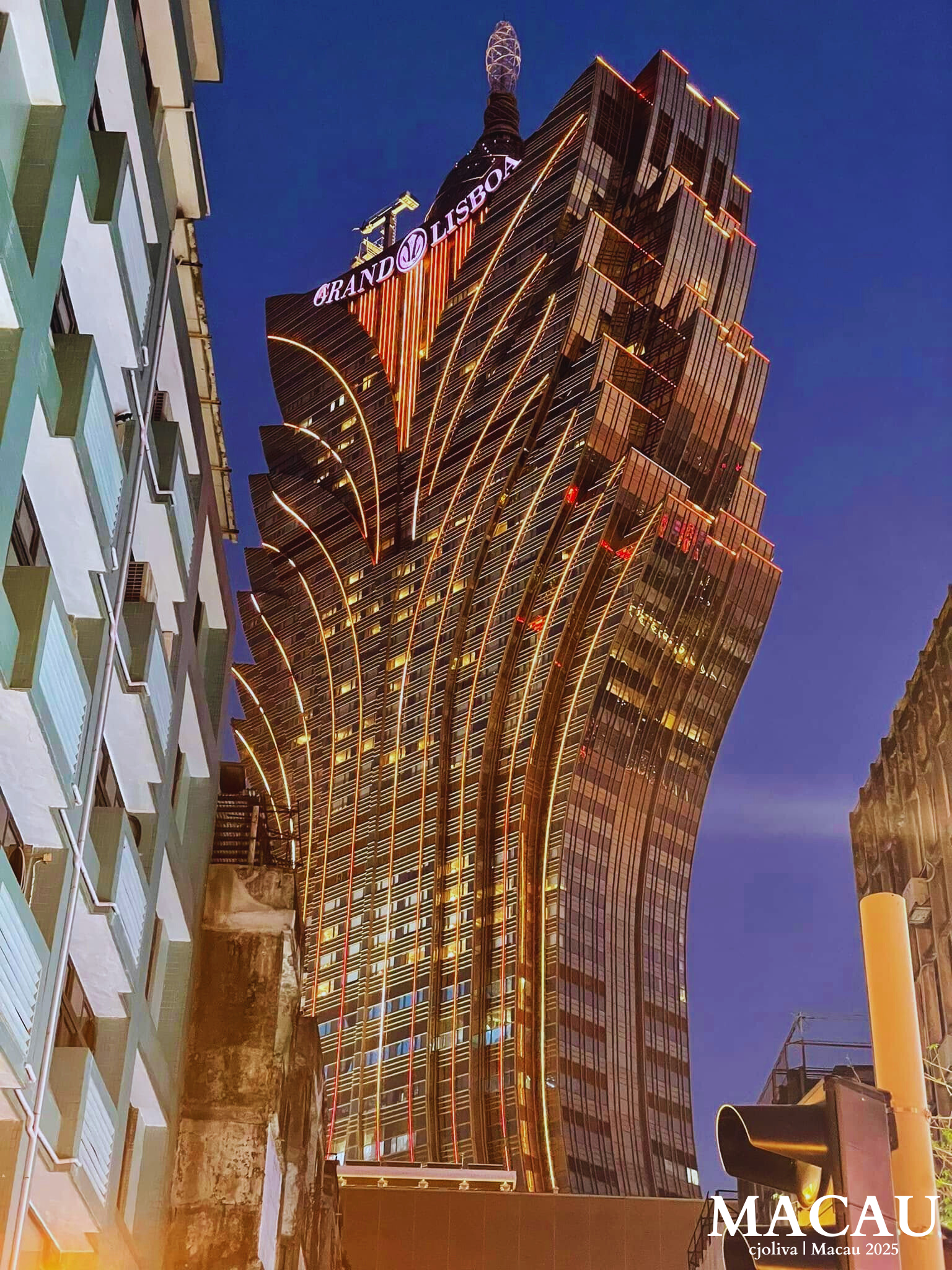 A low-angle nighttime shot of the Grand Lisboa hotel in Macau. The skyscraper's unique lotus-shaped architecture is illuminated in bright gold lights against a deep blue dusk sky, framed by older apartment buildings in the foreground.