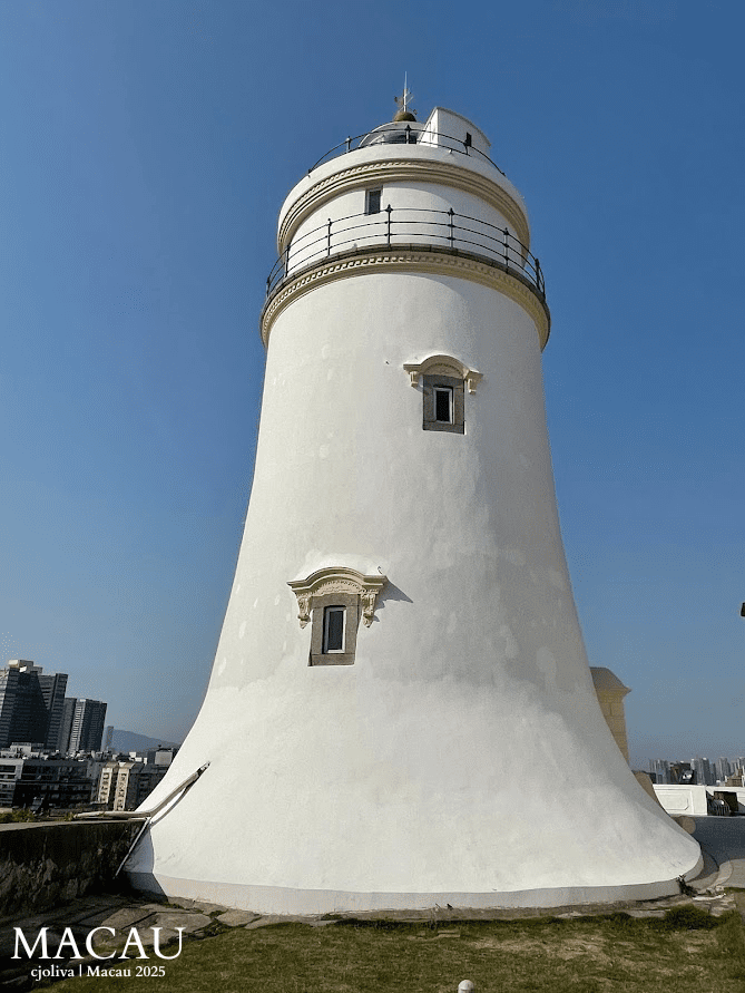 detailed vertical shot of the white Guia Lighthouse with its red dome and black railing, set against a clear blue sky with the Macau city skyline visible in the far background.
