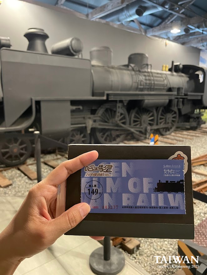 Hand holding a blue admission ticket in front of a large black vintage steam locomotive inside the Hamasen Museum of Taiwan Railway