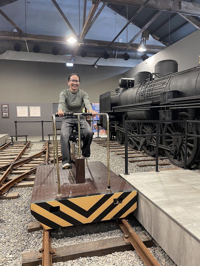 CJ Oliva smiling while sitting on a manual rail bike next to a large black steam locomotive at the Hamasen Museum of Taiwan Railway.