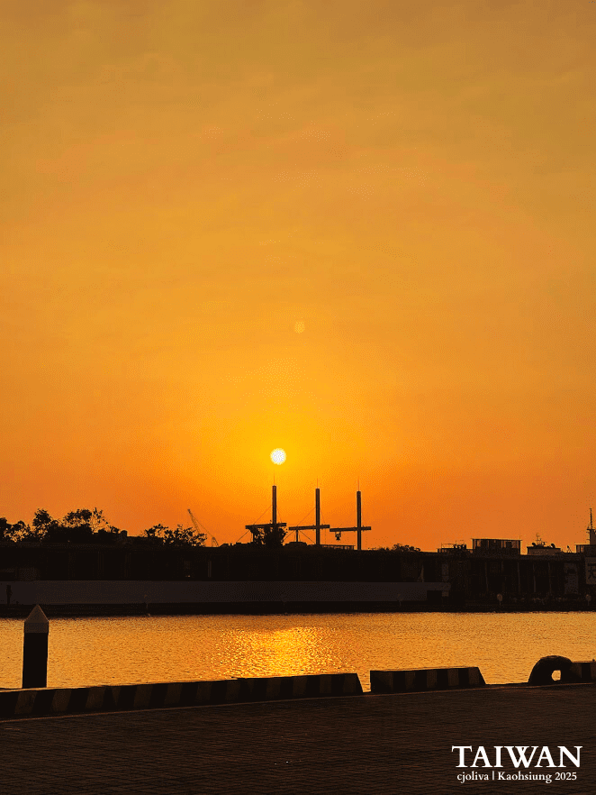 A vibrant orange sunset over the Kaohsiung harbor, with the sun perfectly centered above three tall industrial masts and its golden reflection shimmering on the water's surface.