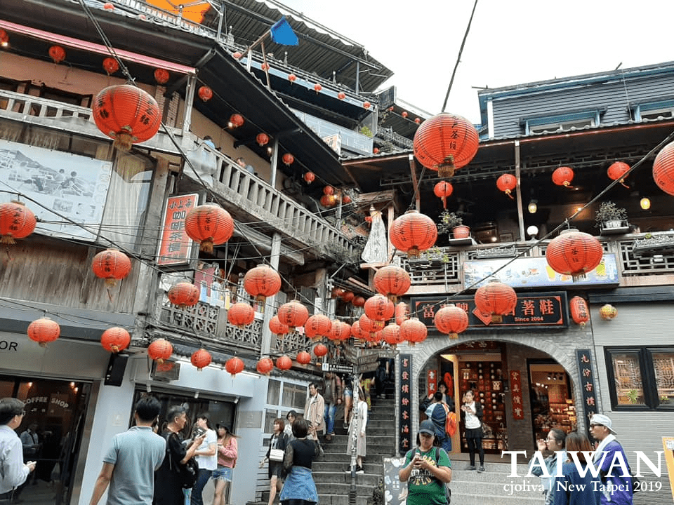 Bustling Jiufen street in New Taipei, Taiwan, with red lanterns hanging across traditional wooden and stone buildings, tourists exploring shops, taken in 2019