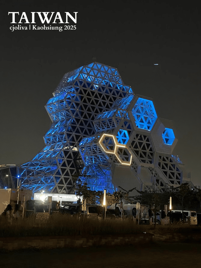 The Kaohsiung Music Center at night, showing its white honeycomb-patterned geometric structure illuminated with bright blue lights against a dark sky.