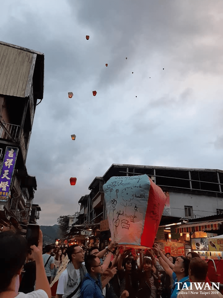 People releasing decorated sky lanterns with messages of luck, health, and success along Shifen Old Street in New Taipei, Taiwan, with multiple lanterns floating into the cloudy sky, taken in 2019