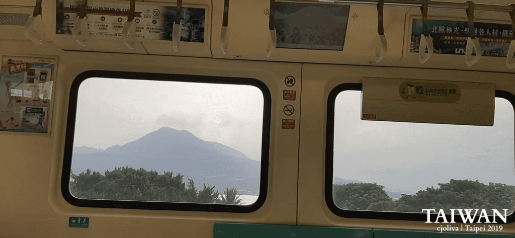 View from inside a Taipei MRT train carriage showing Guanyin Mountain through large windows. The interior features overhead hand straps, transit advertisements, and "No Smoking" or "No Food" stickers, while the outside view shows lush green trees and a misty mountain peak.