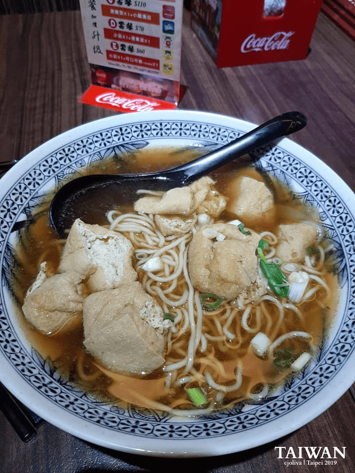Bowl of Taiwanese noodle soup with tofu, green onions, and dark broth on a wooden table in Taipei.