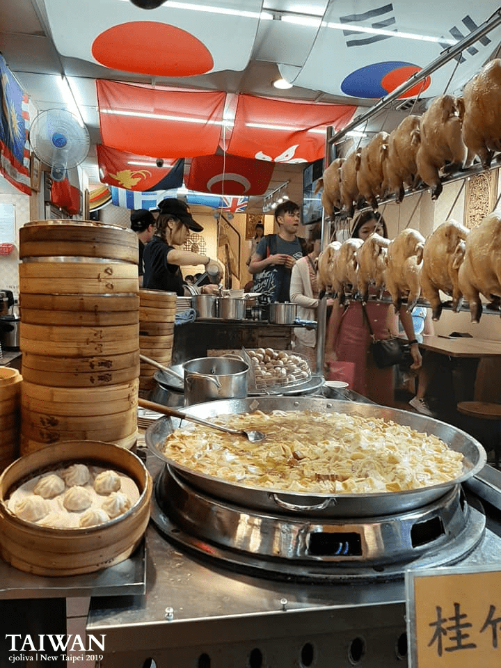 A bustling food stall in Jiufen, Taiwan, with bamboo steamers of dumplings, stir-fried tofu and vegetables, and whole chickens hanging, surrounded by workers and international flags, taken in 2019