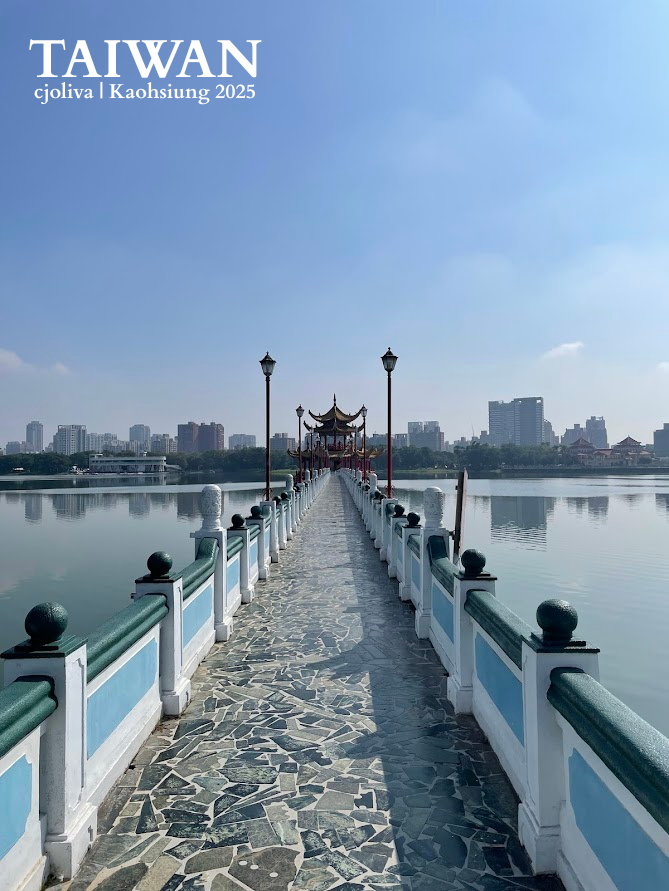 A long, symmetrical stone bridge with blue and white railings leading to a traditional Chinese pavilion on a calm lake in Kaohsiung, Taiwan.