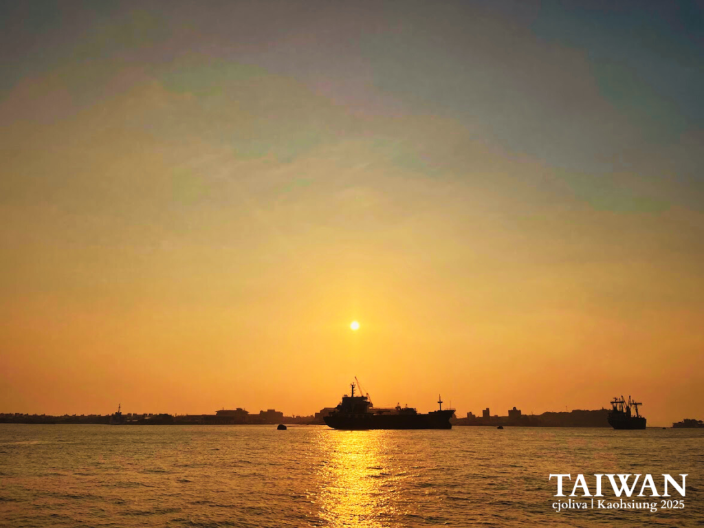 A wide-angle shot of a cargo ship silhouetted against a vibrant orange and yellow sunset in the Kaohsiung harbor, with the city skyline visible on the horizon.