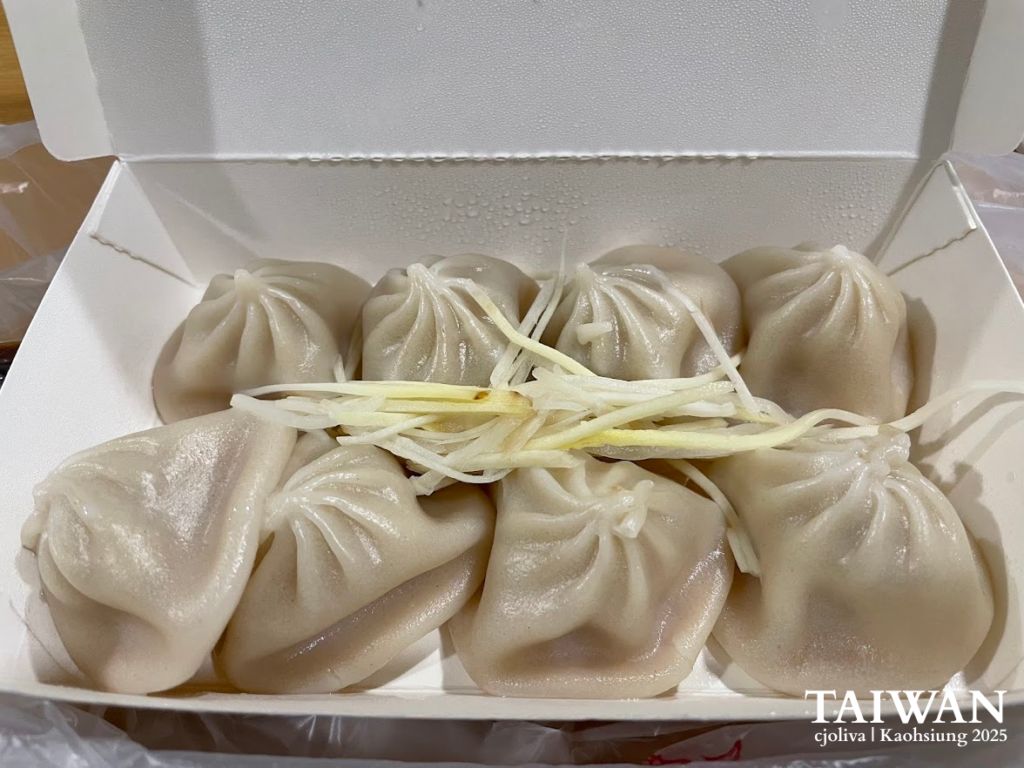 A close-up shot of eight steamed soup dumplings (Xiao Long Bao) in a white takeout box, topped with fresh shredded ginger.